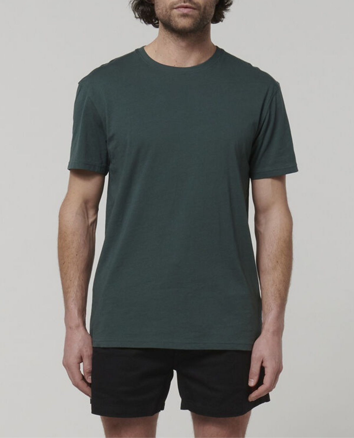A person with medium-length dark hair is wearing a Hard Yakka Core Tee in plain dark green, made from 100% Australian Cotton, accompanied by black shorts. The individual stands against a light gray background, capturing only the upper half of their legs without including the full head.