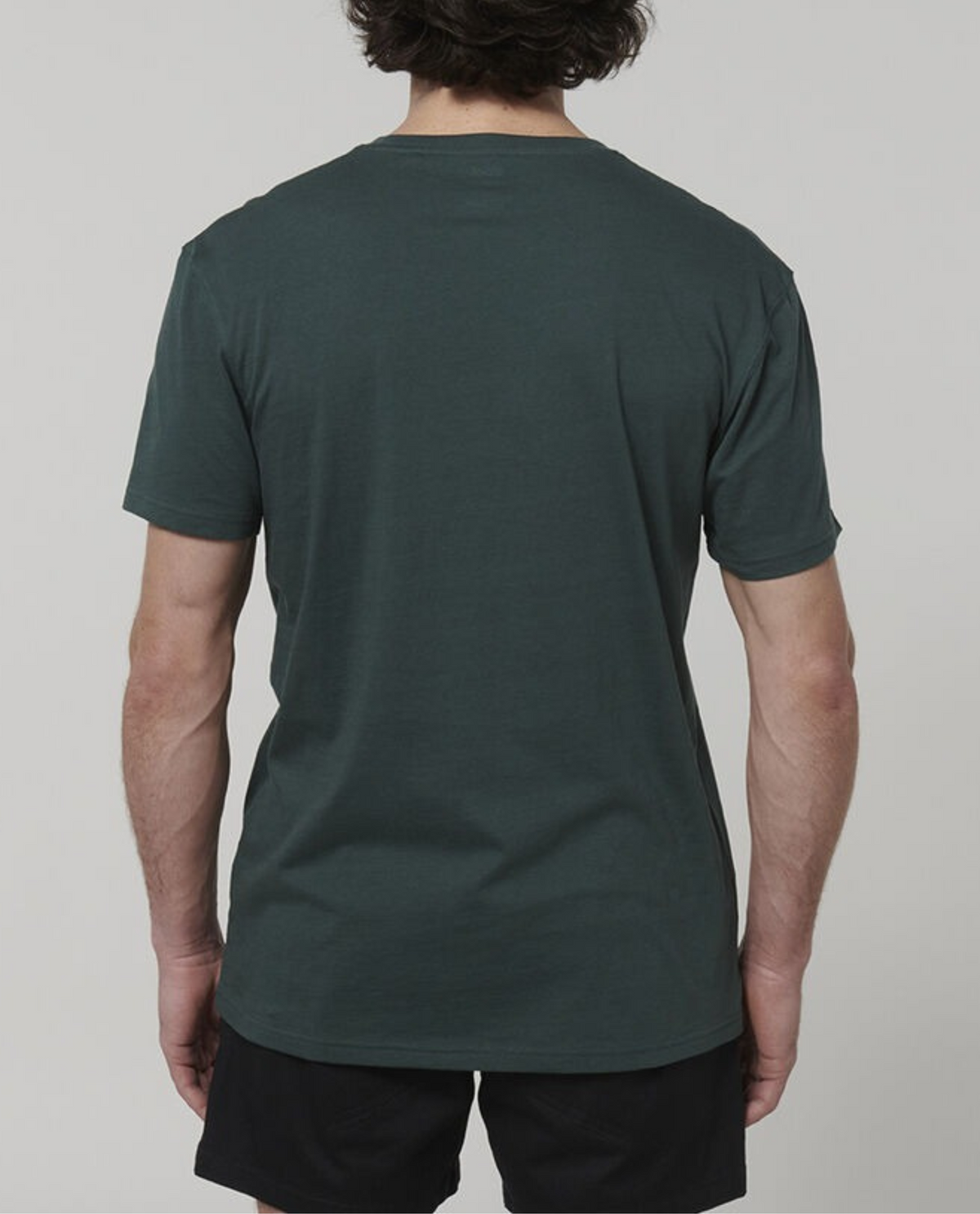 A person with short, dark hair is shown from the back, wearing a Hard Yakka Core Tee made of 100% Australian Cotton in dark green and black shorts. The image background is plain and light-colored, capturing a casual summer work vibe.