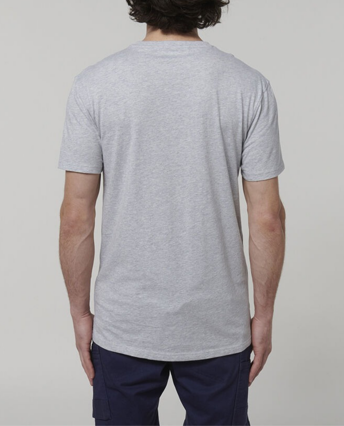 A person with shoulder-length dark hair is standing with their back to the camera, wearing a plain, light gray Hard Yakka Core Tee and dark blue pants against a plain, light-colored background. The tee is made from 100% cotton.