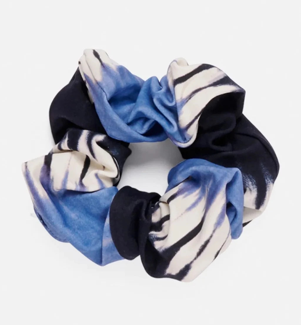 A close-up of a Rip Curl Surf Treehouse Scrunchie with a tie-dye pattern in black, blue, and white colors, resting on a plain white background. The fabric appears soft and gathered, forming the classic circular shape of a hair scrunchie.