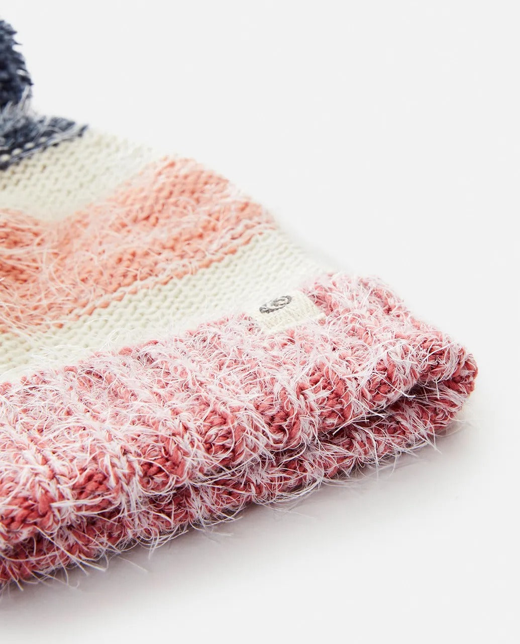 A close-up of the Rip Curl Surf Tree House Pom Pom Beanie features pink, white, and gray horizontal stripes. This stylish accessory has a ribbed texture with a soft, fluffy appearance, ideal for any cozy outfit.