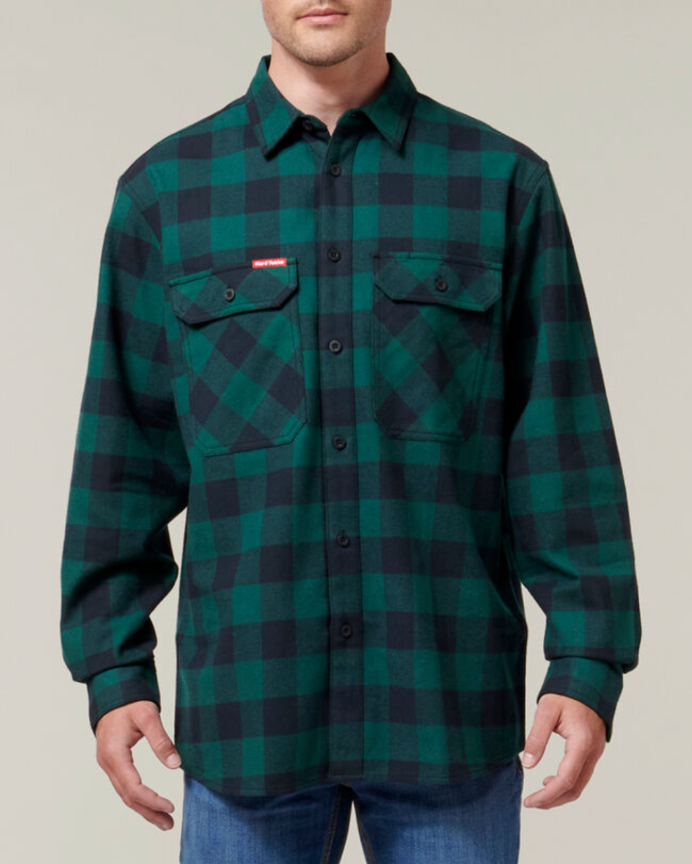 A person wearing the Hard Yakka Check Flannelette Shirt—a durable green and black plaid button-up with two chest pockets—and blue jeans, stands against a plain light background.