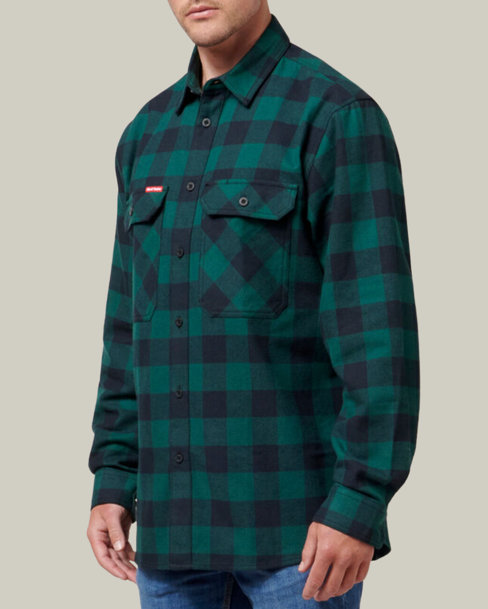 A man in a Hard Yakka Check Flannelette Shirt, featuring durable green and black plaid cotton twill with buttoned chest pockets, and blue jeans, stands against a plain light gray background.