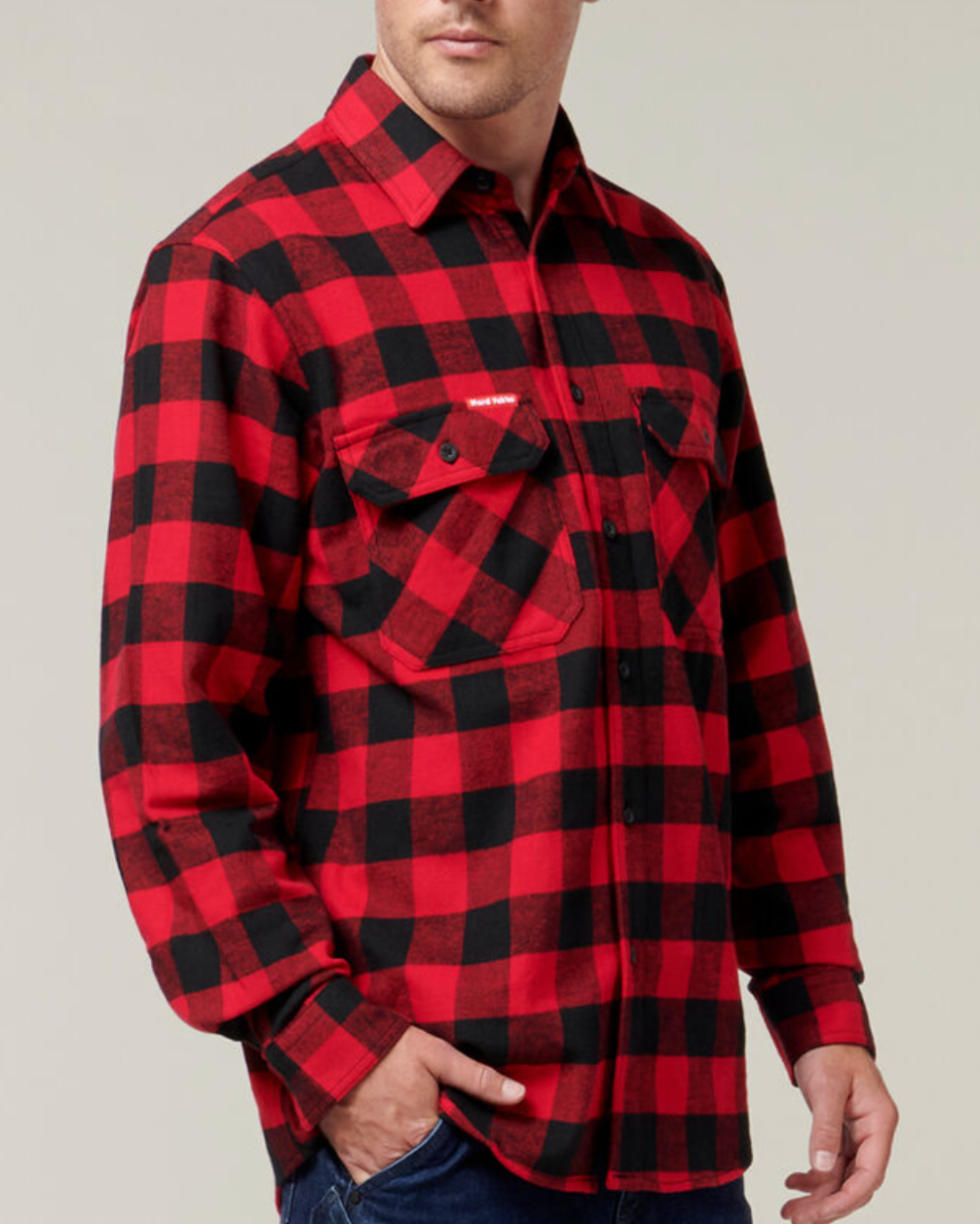 A person wears a Hard Yakka Check Flannelette Shirt in red and black plaid, featuring long sleeves, durable cotton twill fabric, and two chest pockets. They pair it with dark blue jeans against a neutral gray background.