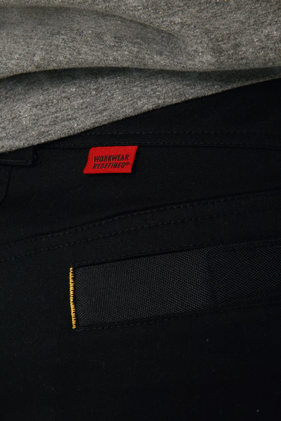 Close-up image showing the waistband of a person wearing black CAT Elite Operator Pant and a grey shirt. The stretch work pant features multiple utility pockets, a red tag with the text "WORKWEAR REDEFINED," and a black belt loop with yellow stitching on the seam.