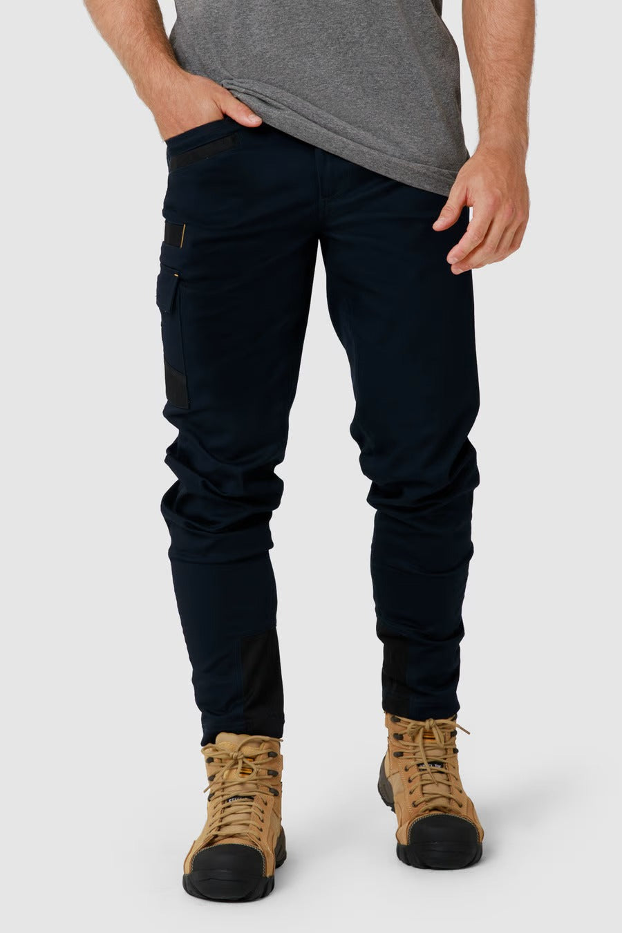 A person in a gray t-shirt and CAT Elite Operator Pant in dark blue, featuring multiple utility pockets, stands with one hand in the pocket. They are wearing tan work boots and are positioned against a plain light gray background.