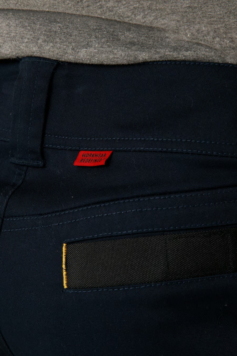 Close-up of the back pocket of a pair of CAT Elite Operator Pant in navy blue stretch fabric, showcasing a small red tag with black text and a black detail on the pocket. The top edge of a grey shirt is visible above the slim tapered fit trousers.