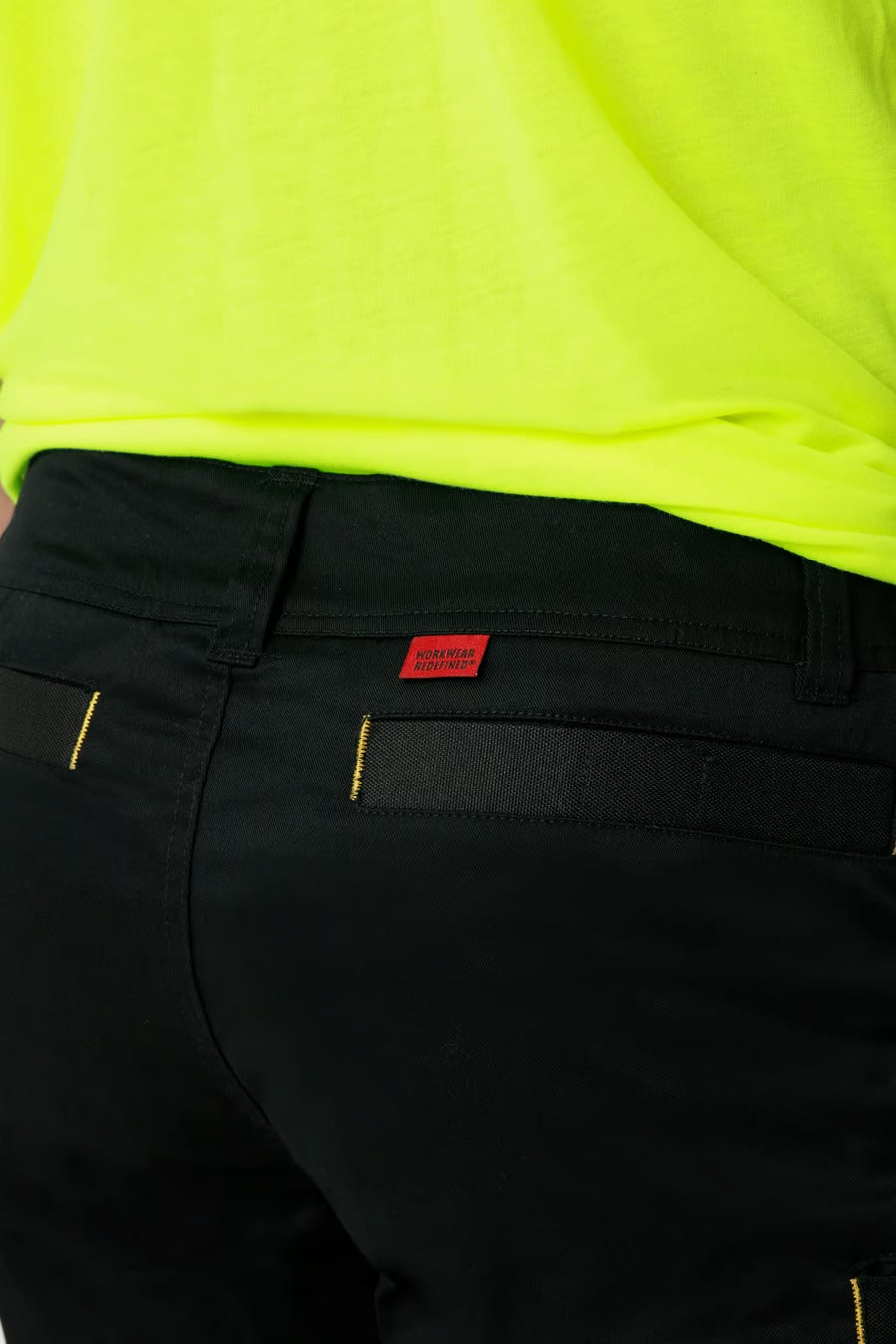 A close-up image features a person wearing the CAT Elite Operator Short in black, showcasing its utility pockets. The individual is also sporting a bright yellow top. A small red tag with text is visible at the back of the waistband on the shorts, though it is not fully readable in the image.
