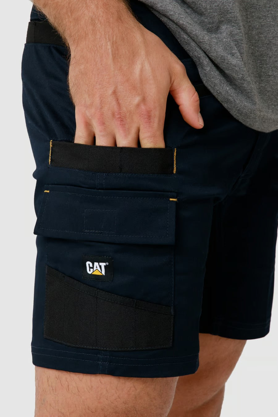 A person wearing a pair of dark blue CAT Elite Operator Shorts is shown. The shorts feature multiple utility pockets, including a velcro flap pocket with the CAT logo and an additional smaller pocket above it. The individual, dressed in a grey shirt, is placing their hand into one of the pockets.