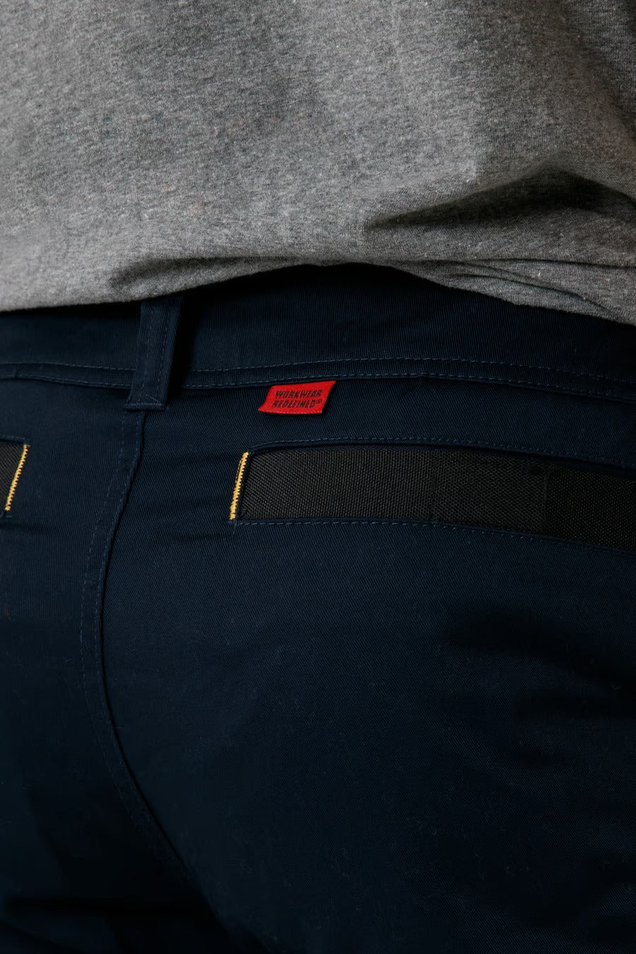 A close-up image showcasing the dark blue CAT Elite Operator Short, featuring yellow stitching on the utility pocket and a small red tag above it. The person is also wearing a gray shirt.