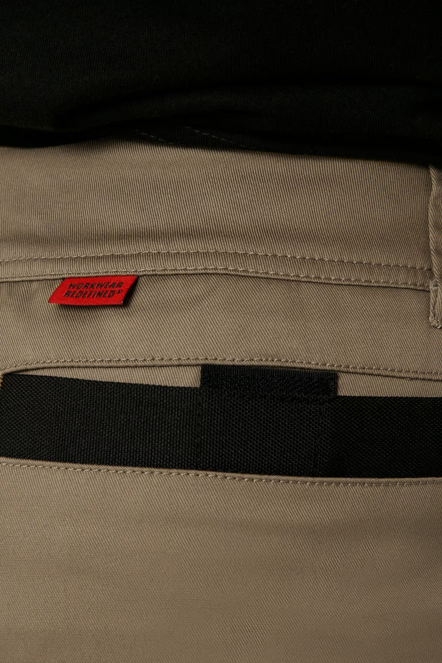 Close-up image of the back pocket of a pair of CAT Elite Operator Shorts in khaki stretch fabric, featuring black details. A small red tag near the pocket seam displays "WISCONSIN LIGHTHOUSE." The waistband, elastic inserts, and belt loops are also visible.