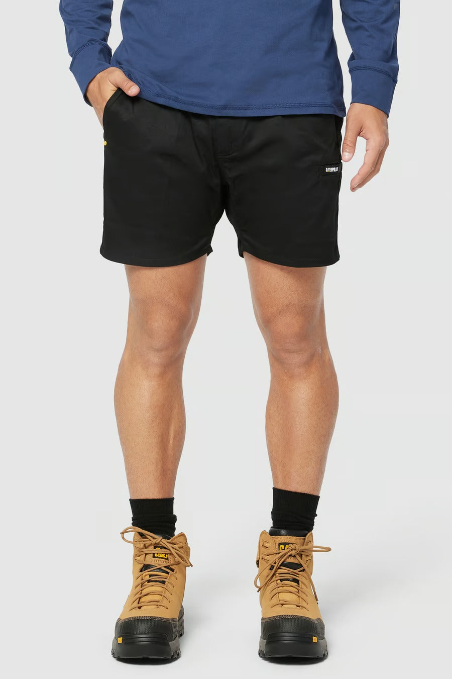 A person modeling the CAT Short Haul Short, featuring a small white logo on the left leg, paired with a blue long-sleeve shirt. The individual is also wearing black socks and beige hiking boots, standing against a plain light background.