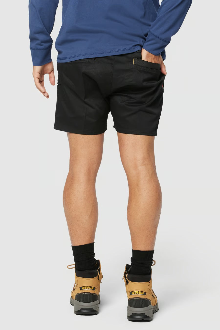 Back view of a person wearing a long-sleeve blue shirt, black CAT Short Haul Shorts with an elastic waist, black socks, and tan hiking boots. The person is standing with hands in their back pockets against a plain white background.