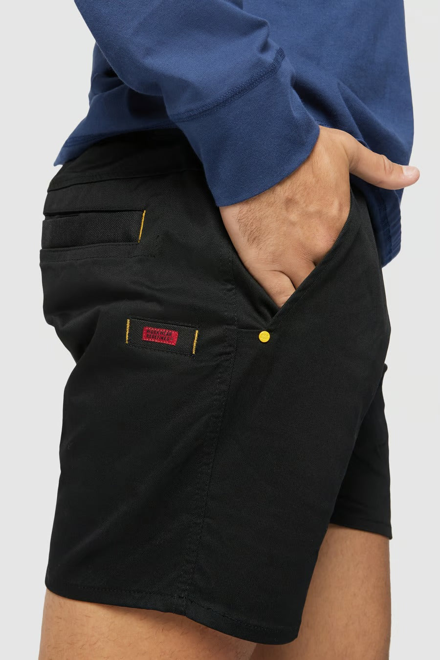 A person wearing black CAT Short Haul Shorts with an elastic waist and pockets, paired with a blue long-sleeve shirt, has their right hand in the pocket. The detailing on the shorts includes yellow stitching and a small red label on the upper part of the pocket.