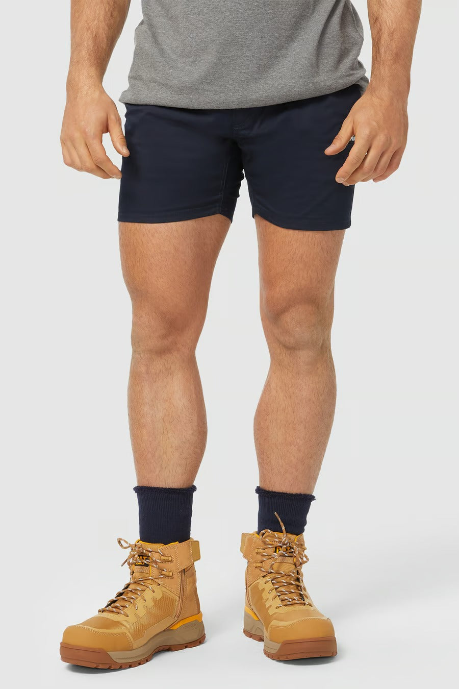 Below the waist, a person stands against a plain background wearing navy blue CAT Short Haul Shorts with an elastic waist and a cargo pocket. They pair these shorts with black socks and tan work boots, although only the lower half of their body is visible.