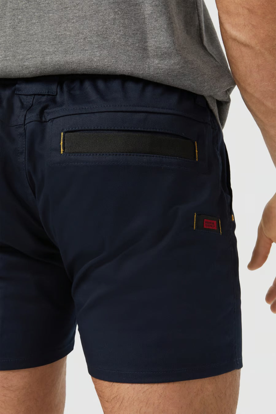 A person wearing the CAT Short Haul Short in navy blue and a grey shirt is shown. A close-up image features the back cargo pocket of the shorts, which has a flap secured by a black strip and yellow stitching. The fabric tag beside the pocket is visible with red and black details.