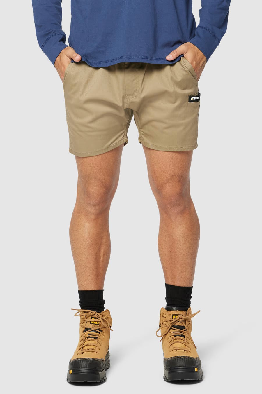 A person wearing a blue long-sleeved shirt, CAT Short Haul Short in beige with cargo pockets and hands in pockets, black socks, and tan lace-up boots. The background is plain white.
