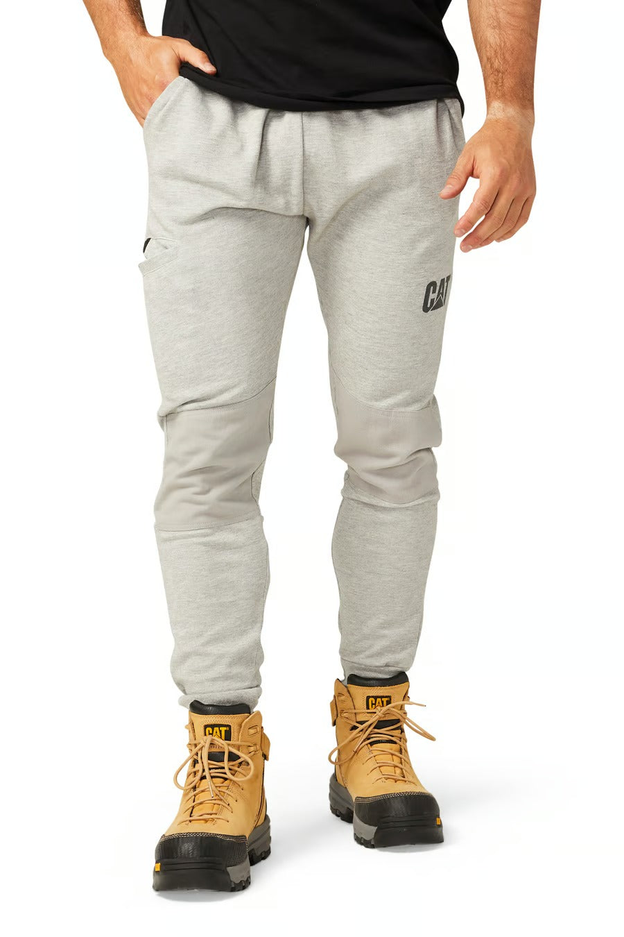 A person stands against a white background, wearing CAT Track Pants in grey with "CAT" lettering, a black shirt, and tan work boots featuring durable black soles.