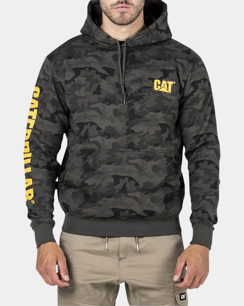 Wearing a CAT Trademark Banner Hooded Sweat with a camouflage pattern and yellow "CAT" logos on the chest and left sleeve, a person stands against a white backdrop. They pair it with beige cargo pants.