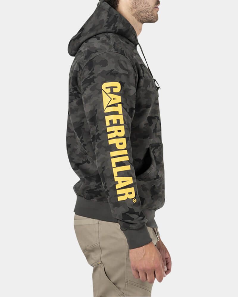 A person stands in profile wearing the CAT Trademark Banner Hooded Sweat, showcasing a camo pattern and bold yellow CAT logo on the sleeve. The hoodie has a cozy fleece lining and is paired with beige pants against a plain white background.