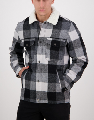 A person in a stylish Swanndri Kaituna Sherpa Lined Jacket stands with hands in pockets. The checkered jacket features a plush sherpa lining, a white fluffy collar, and buttoned front pockets against a plain white background.