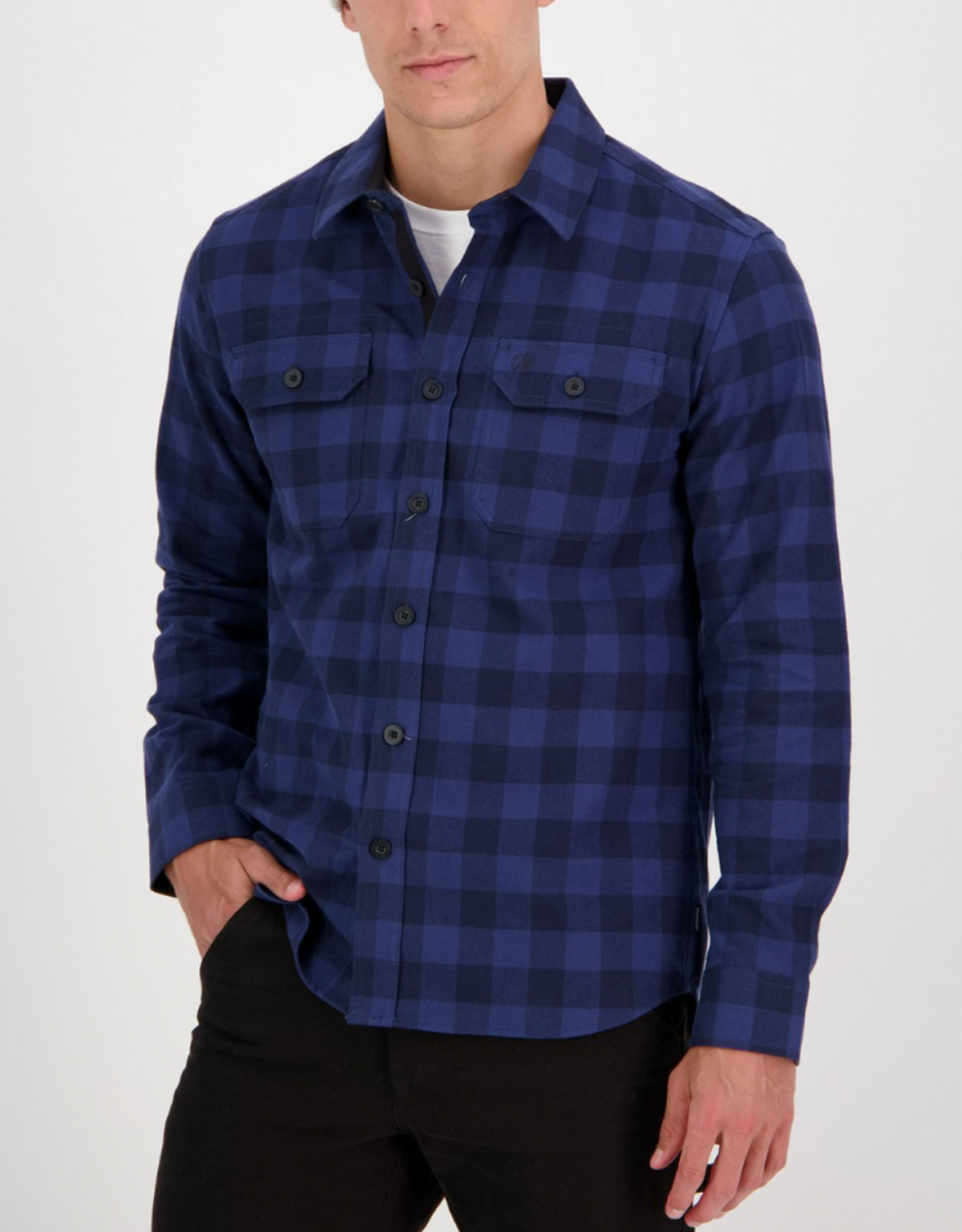 A man wears the Swanndri Taranaki Tailor Shirt—a stylish, 100% cotton blue and black checkered button-up—over a white t-shirt, with his left hand in his pocket, standing against a plain light background.