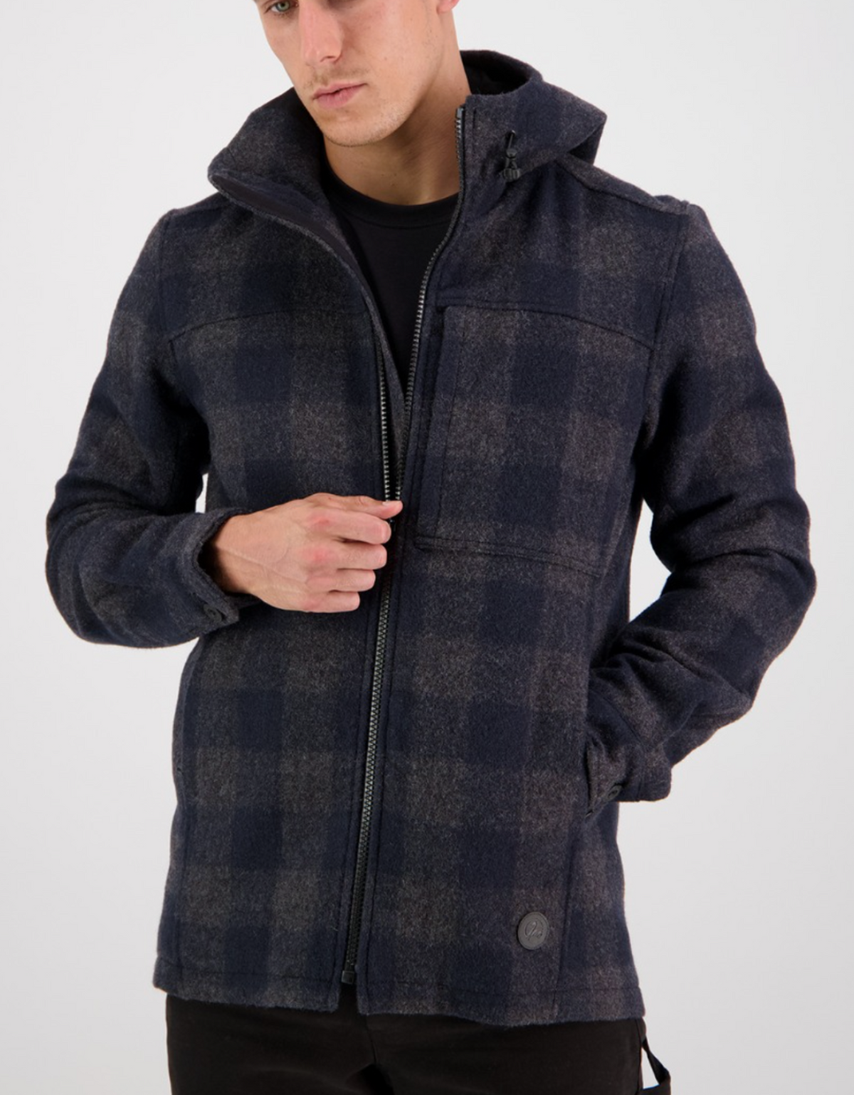 A person stands against a plain background wearing a Swanndri Hudson Hoody, featuring a dark navy checkered pattern and high collar. Made from New Zealand wool, the hoody is partially zipped over a black shirt.