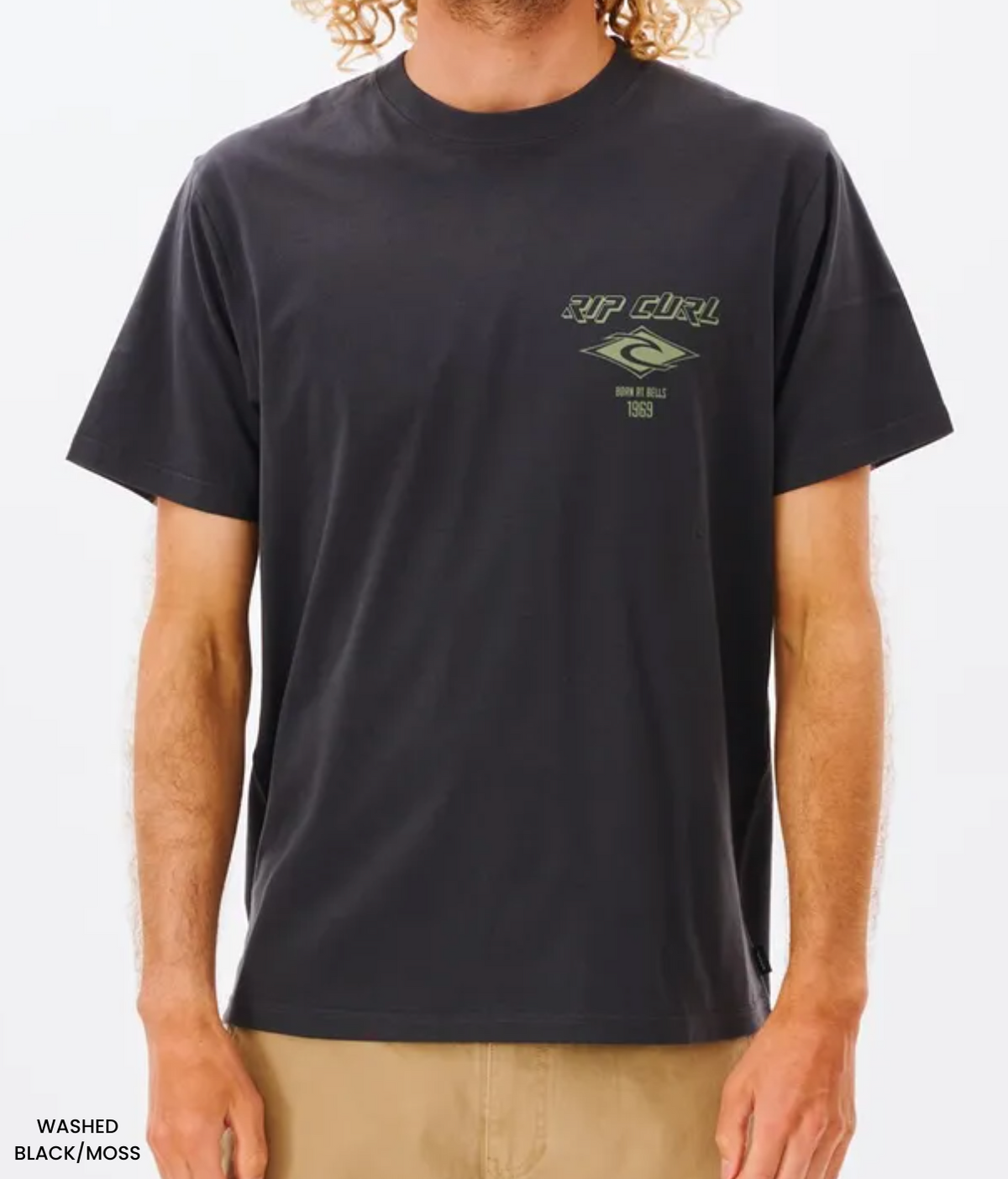 A person wears the black Rip Curl Fade Out Icon Tee in a relaxed fit, featuring an abstract design with "RIP CURL" and "BORN AT BELLS 1969" in green on the chest. Paired with beige pants, it complements their curly hair for an effortlessly cool look.