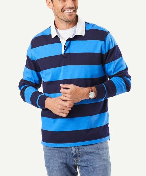 A man wearing the Gazman Block Stripe Rugby, a long-sleeve, regular fit shirt with blue and navy stripes, and light blue jeans is standing against a plain background. He is smiling and holding his hands together. The cotton shirt features a white collar, and the man is wearing a wristwatch.