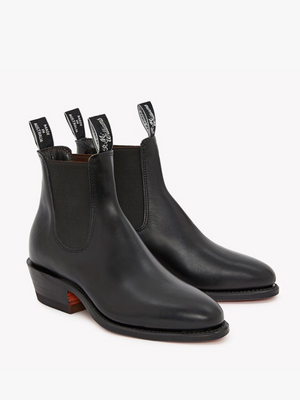 The RM Williams Lady Yearling Boot is a black leather pair with elastic side panels, pull tabs with white text, low stacked wooden heels, and rounded toes, shown against a white background.