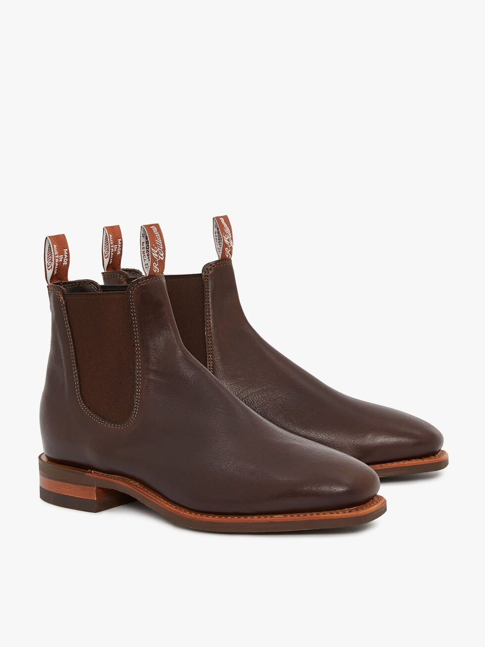 The RM Williams Comfort Kangaroo Craftsman Boot is a pair of handcrafted brown leather Chelsea boots made in Australia. They feature pull tabs with branded tags, elastic side panels, a low stacked heel, and a smooth finish. The boots are shown to display both the outer and inner sides.