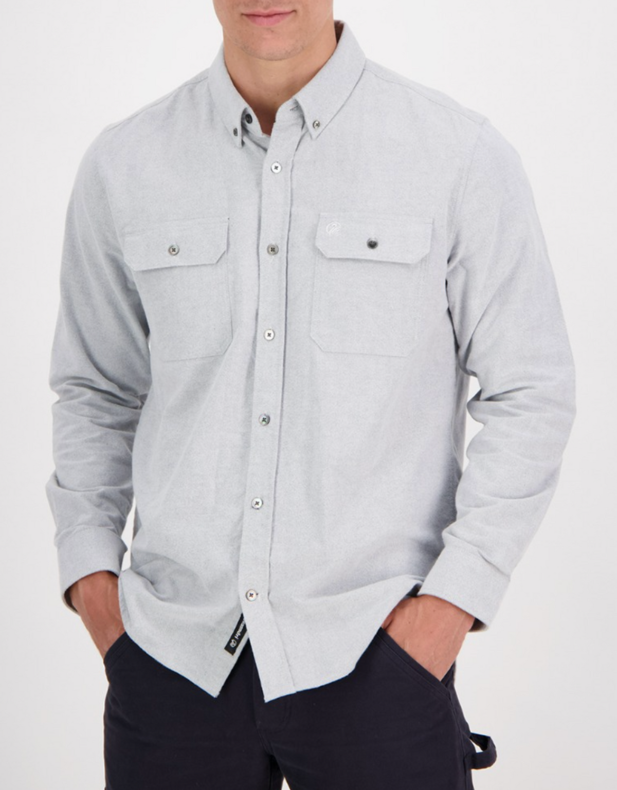 A person sports a Swanndri Marwood Shirt in light gray, a stylish classic-fit button-up made of 100% cotton. It features two chest pockets, and their hands rest casually in the pockets of their dark pants against a plain white background.
