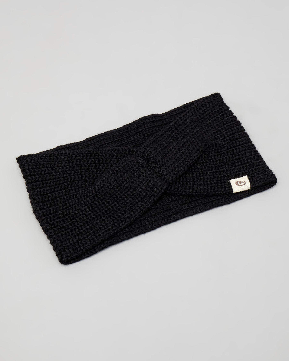 The Rip Curl Coco Headband, a black cotton knit with a twisted design, is displayed on a light gray background with a small white label featuring the Rip Curl logo attached at the edge.