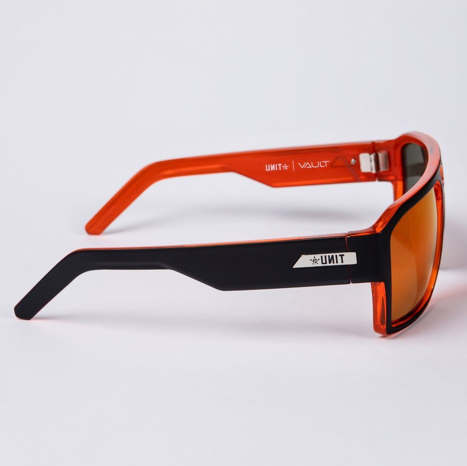 A pair of UNIT Vault Polarised Eyewear featuring an orange frame, black arms, and orange-tinted lenses. The left arm displays a silver logo near the hinge alongside white text that reads "UNIT," while the interior of the right arm is marked with white text that reads "UNIT | VAULT." These sunglasses offer UV protection and high sun glare protection, all set against a white background.