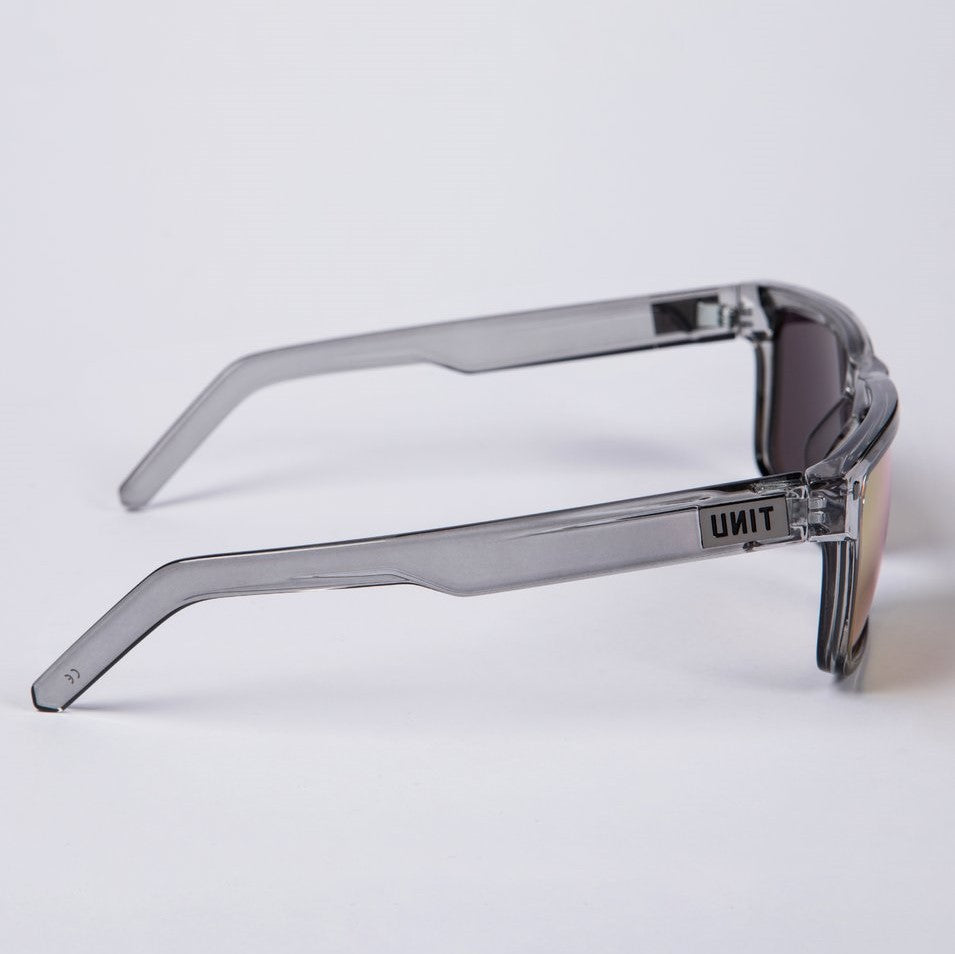 A pair of UNIT Primer Polarised Eyewear, featuring gray-tinted, polarized lenses and transparent gray, lightweight frames, positioned on a white surface. The left temple arm of the sunglasses displays "UNIT" in black capital letters, ensuring both style and UV protection.