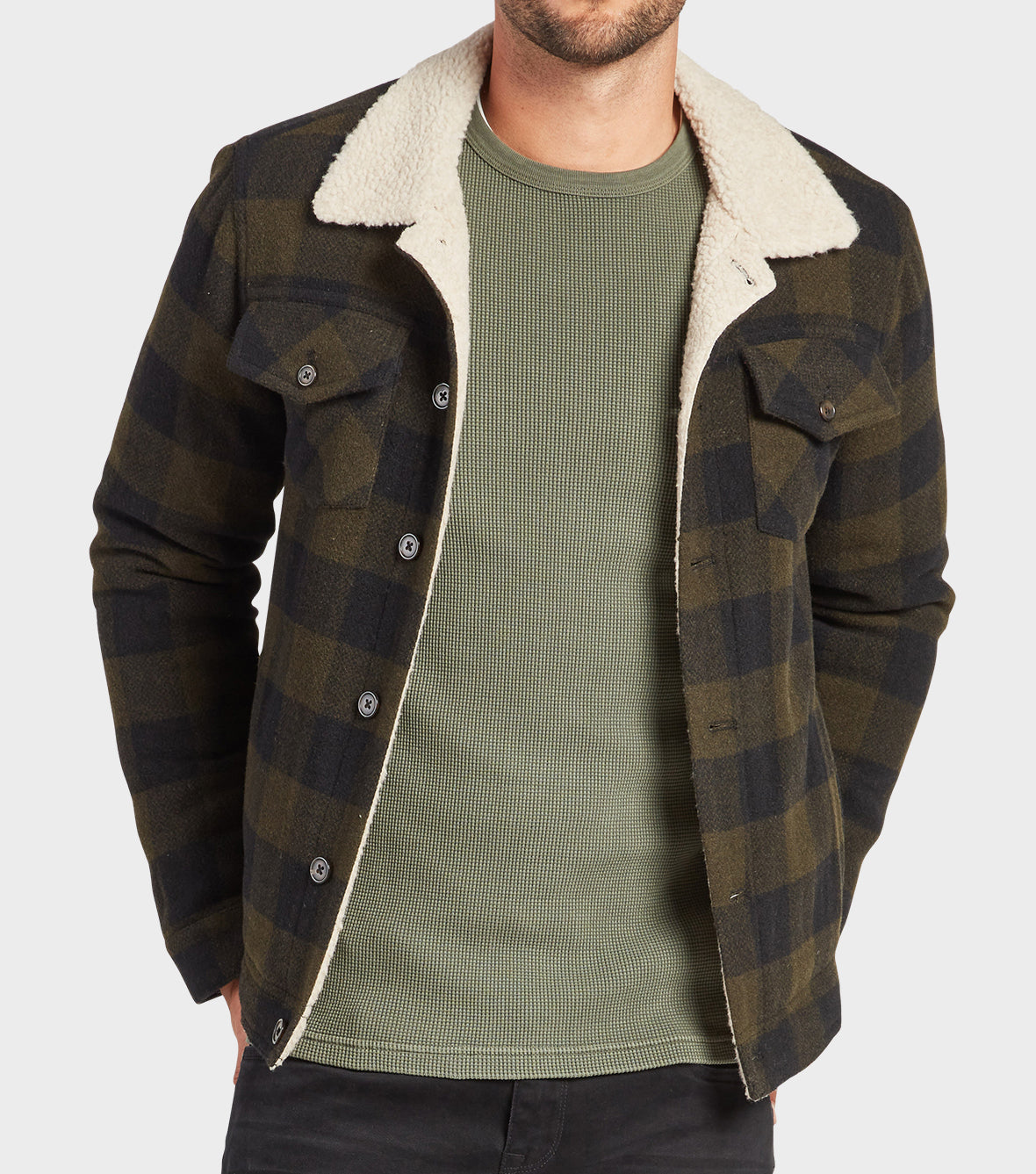 A man in a green waffle-knit shirt and an open Academy Brand Davie Jacket with a white sherpa collar stands with his hands in his pockets against a plain background.