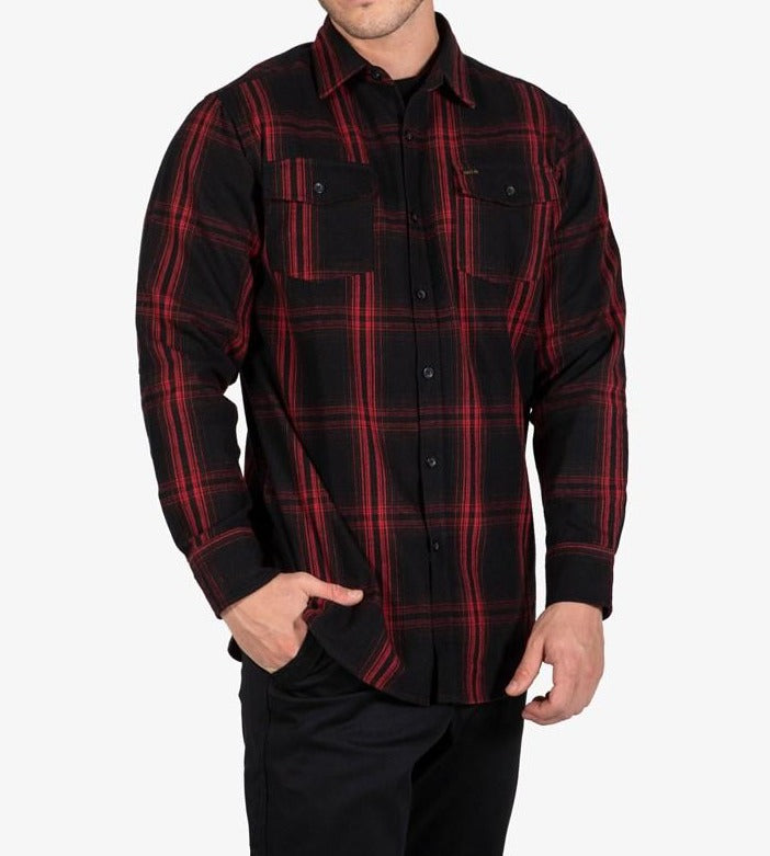 A person is wearing the UNIT Newtown Flannel Shirt, which features a black and red checkered pattern, is made from 100% cotton, and has two buttoned chest pockets along with long sleeves. The individual stands with one hand in their pocket, showcasing the shirt's design against a plain white background.