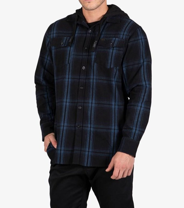 A man stands against a plain white background wearing the UNIT Chester Hood Flannel Shirt in black and blue with a button-up front, paired with black pants and one hand in his pocket.