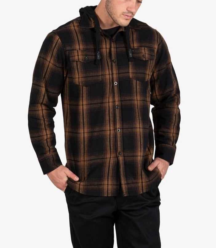 A man wears the UNIT Chester Hood Flannel Shirt, featuring a black and brown plaid design, standing with hands in pockets against a plain light background.