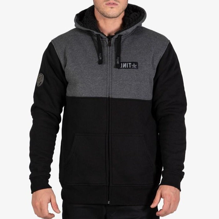 A person wears the UNIT Terrace Fleece Hoodie—two-tone with a dark gray top, black bottom, fleece lining, front pockets, and UNIT logo patches on the left chest and sleeve—standing against a plain light background.