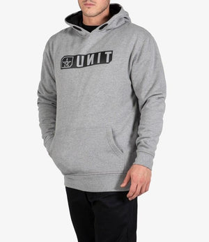 A man stands against a plain white background wearing a comfortable gray UNIT Stride Fleece Hoodie with a front pocket and the black logo printed in reverse on the chest, paired with black pants.