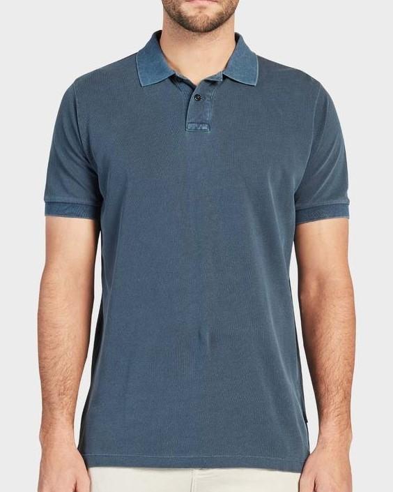 A person is wearing the Academy Brand Academy Polo, a blue short-sleeved, comfortable polo shirt with a collar and three buttons near the neckline. This cotton pique shirt from the Academy Brand has a textured appearance, with sleeves and collar in a slightly darker shade. They are also wearing light-colored pants.