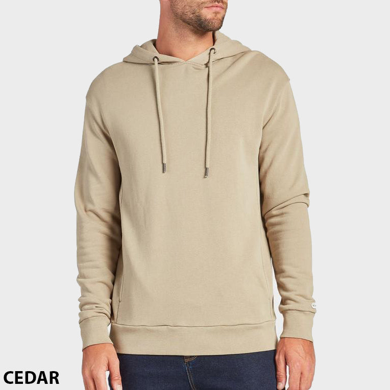 A man stands against a plain background wearing the Academy Brand Boston Hoodie in light beige with drawstrings and dark jeans. "CEDAR" is written in black text at the bottom left corner.