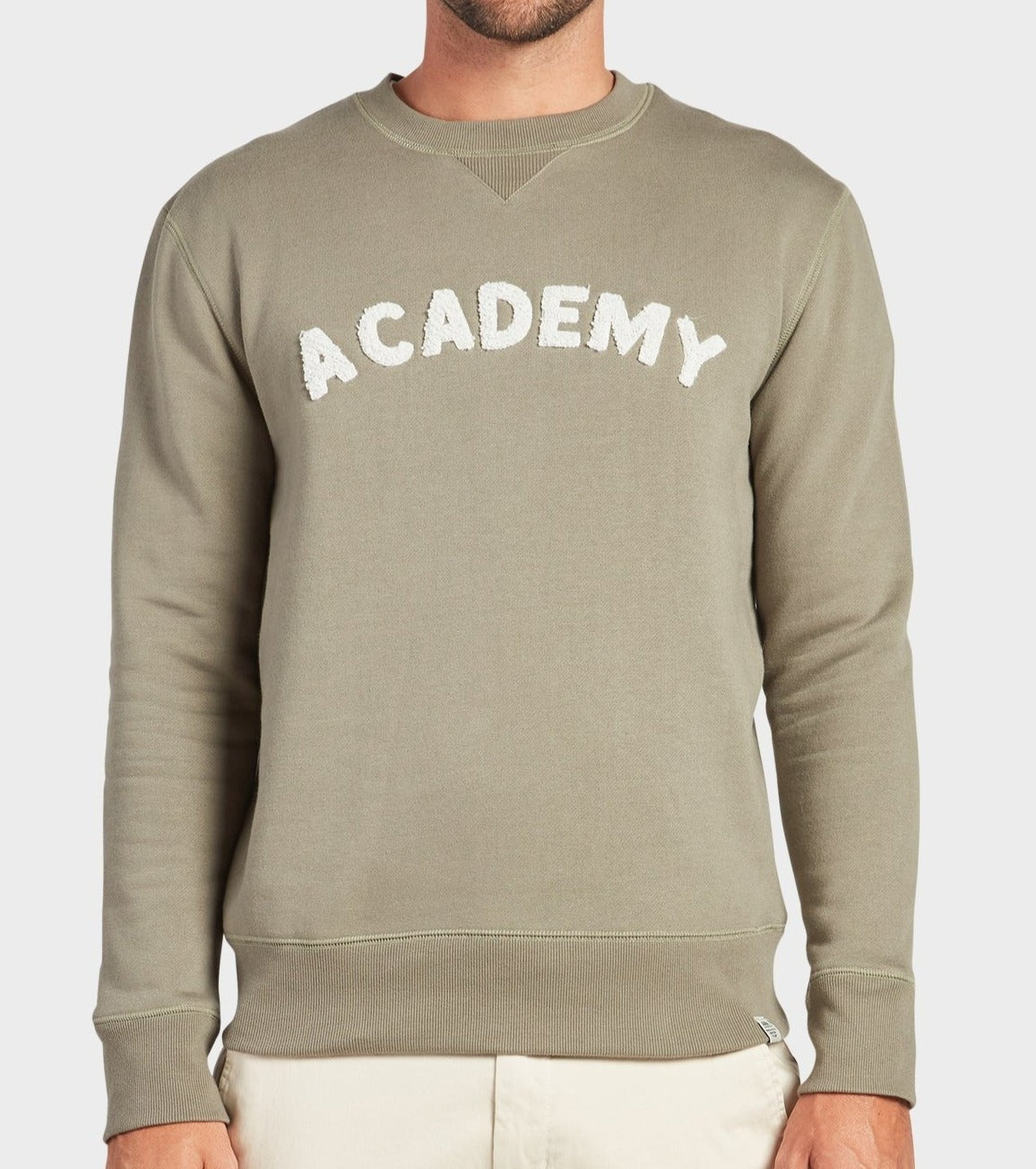 A person wearing the Academy Brand College Crew sweatshirt in Moss, featuring "ACADEMY" in large white letters on the chest and styled casually with light-colored pants. The person's head is not visible.