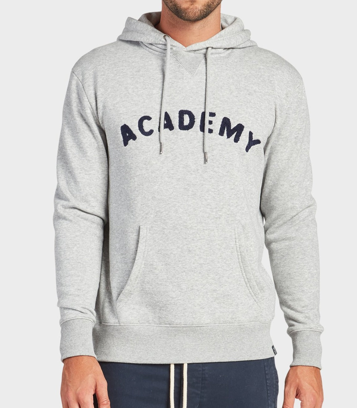 A man wears the Academy Brand College Hoodie in Oxford Heather, featuring "ACADEMY" in bold across the chest, a front pocket, and drawstrings. He pairs the casual sweatshirt with dark pants for a relaxed style.