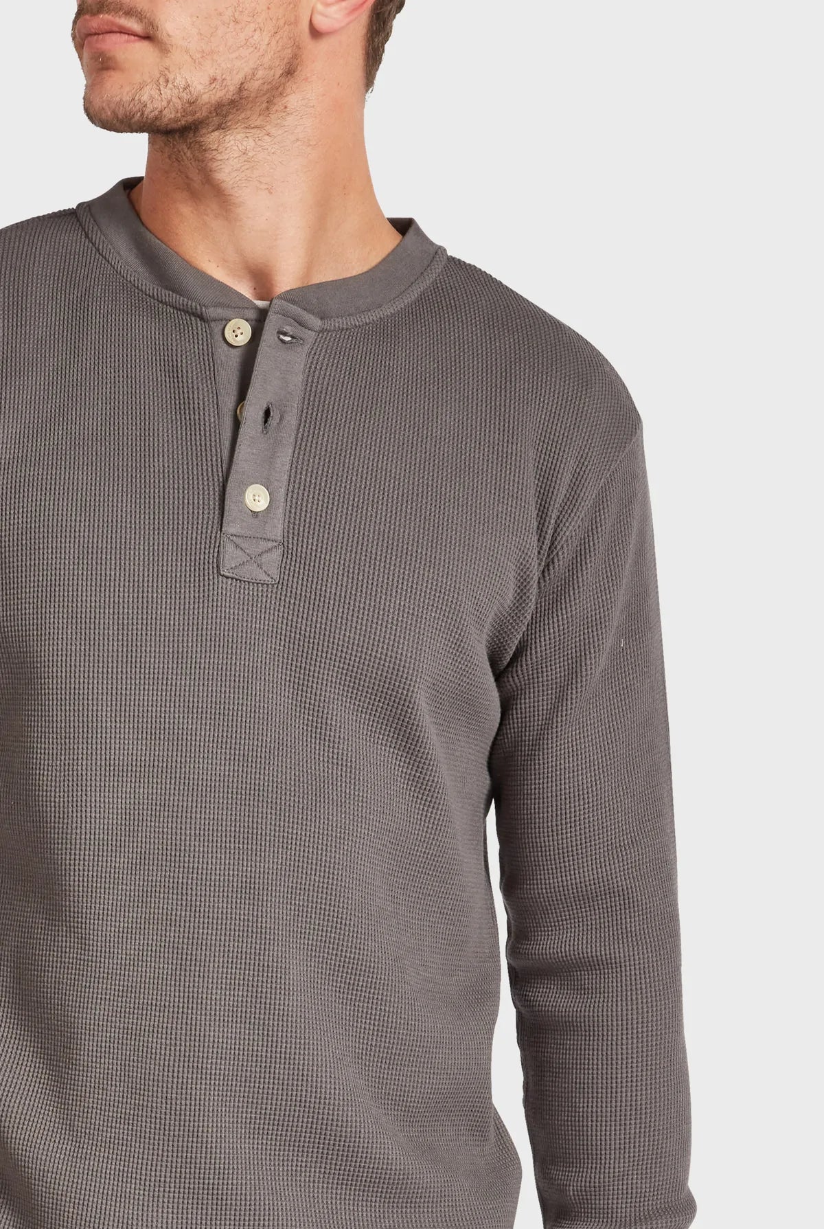 A person wearing the Academy Brand Sycamore Henley, a grey waffle-knit henley shirt made from 100% cotton, with three buttons partially undone. The person is turned slightly to the side, showcasing the shirt's texture and casual style against a plain white background.