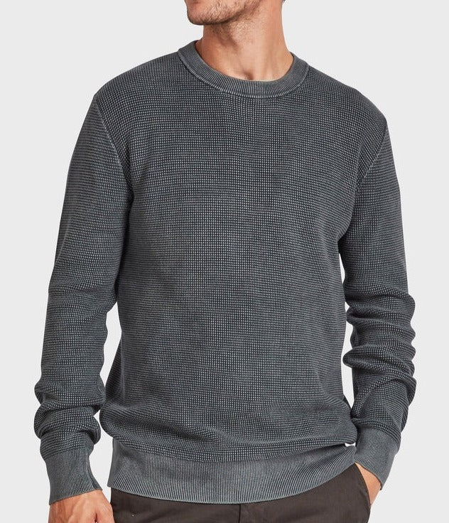 A man in the Academy Brand Newport Crew Knit—a textured dark gray 100% cotton, long-sleeve jumper—stands with one hand in his pocket against a plain light background, head slightly turned and face partially out of frame.