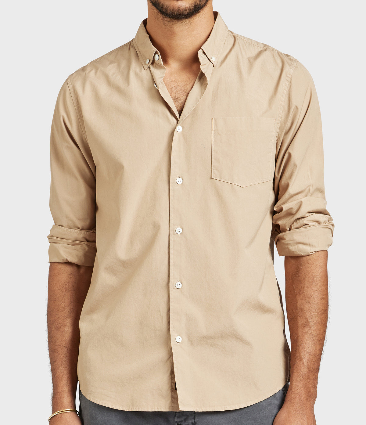 Someone is wearing the Academy Brand Frank Poplin Shirt in beige, made of 100% cotton for comfort, featuring rolled-up sleeves and a chest pocket. It is paired with dark pants against a light grey background.