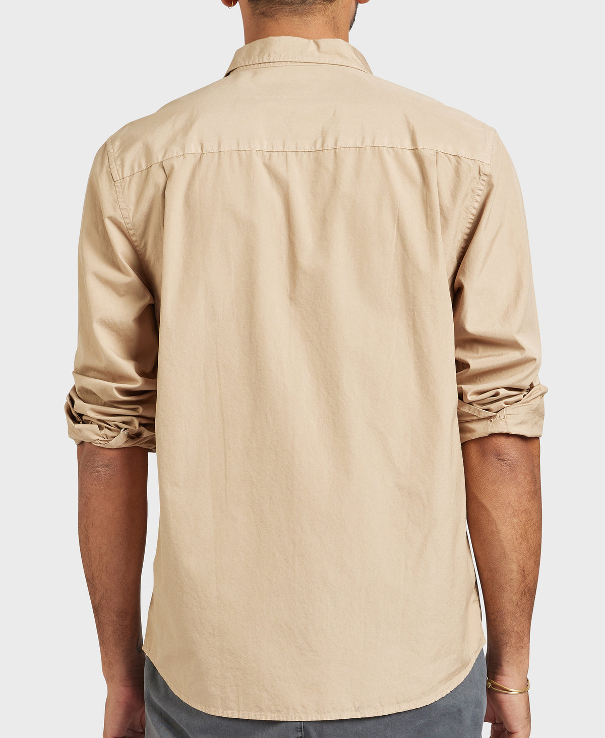 A person sports an Academy Brand Frank Poplin Shirt, a comfortable, light brown, 100% cotton long-sleeve with stylishly rolled-up sleeves. They face away from the camera against a simple white backdrop.