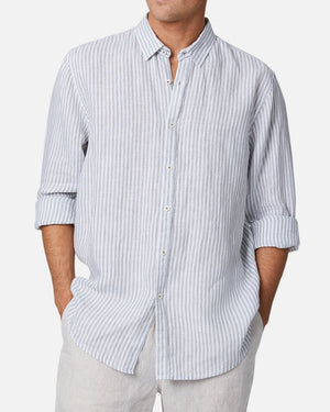 A man wearing the Industrie The Southwest Linen L/S Shirt, featuring light blue and white vertical stripes with rolled-up sleeves, paired with light-colored pants and standing with his hands in his pockets for a relaxed look.